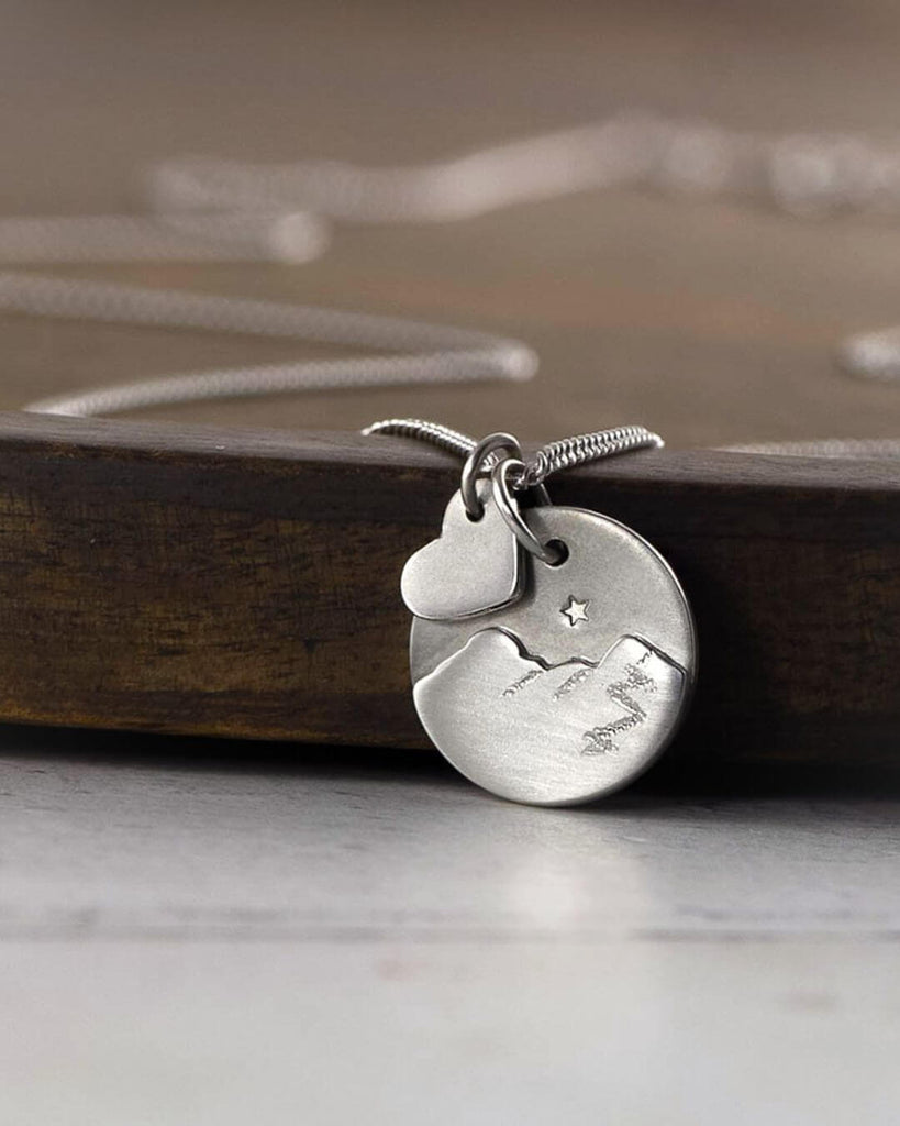 Handmade silver Catbells pendant with a little silver heart resting on a dark board.