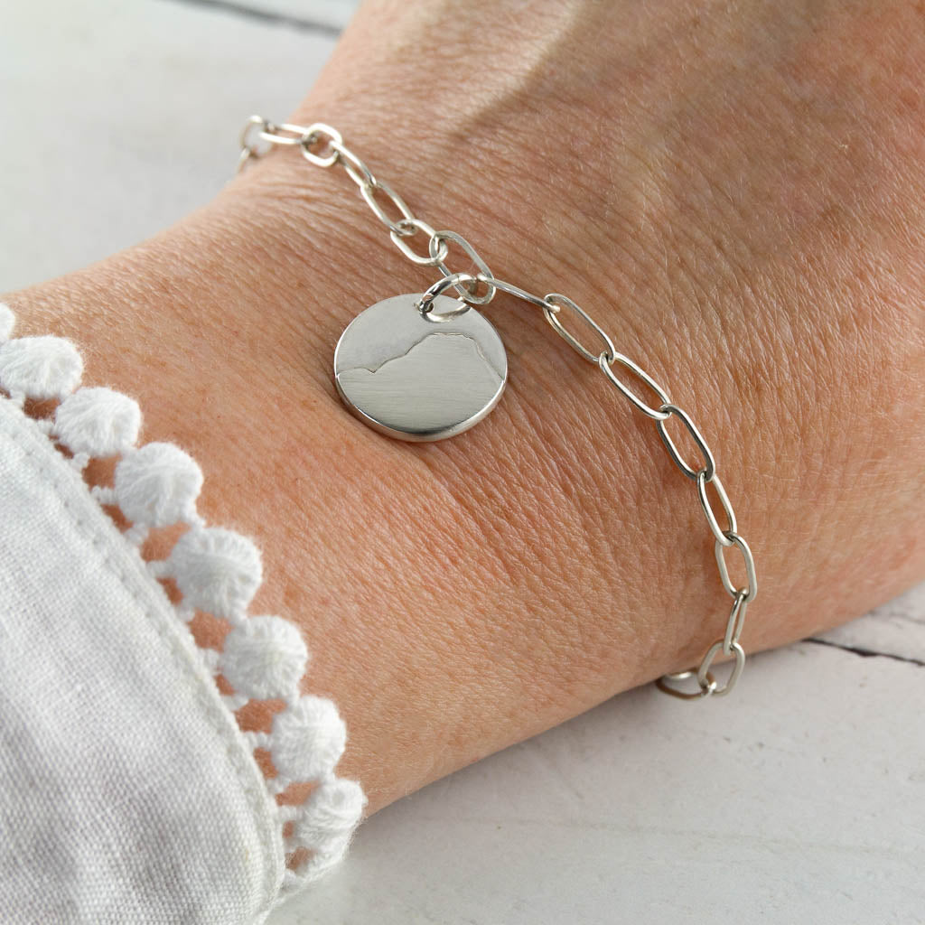 Silver chain bracelet with a round charm of Raven Crag in the Lake District, on a wrist against a light background